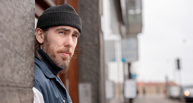 Han står och lutar sig mot en vägg. Han har skägg och en svart mössa.