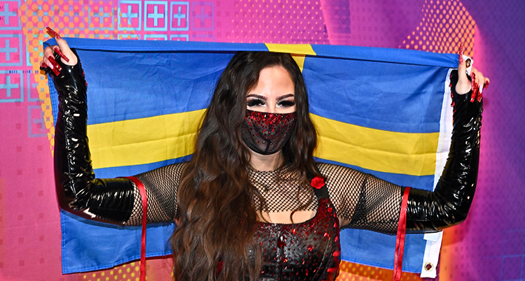 Hon håller upp Sveriges flagga. Hon har ett svart munskydd i spets över munnen.