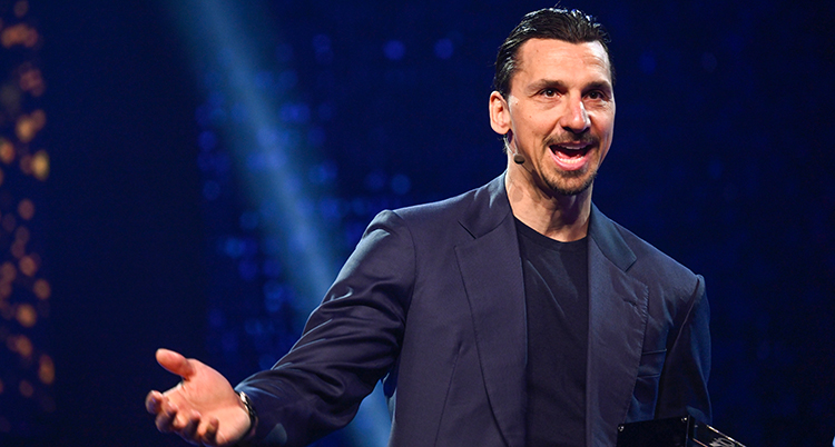 Han har mörk kostym på sig. Han pratar på en scen.