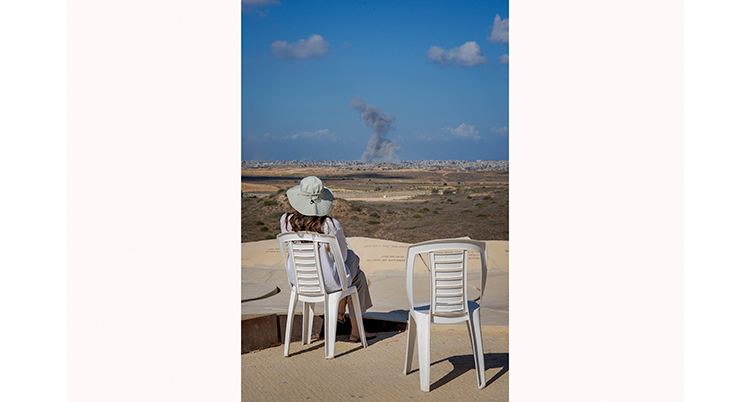 En kvinna i vit hatt sitter och tittar mot horisonten. Där syns en stad och rök. Himlen är blå.