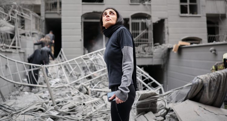 Hon står bland sönderbombade hus. Hon har en mobiltelefon i handen.