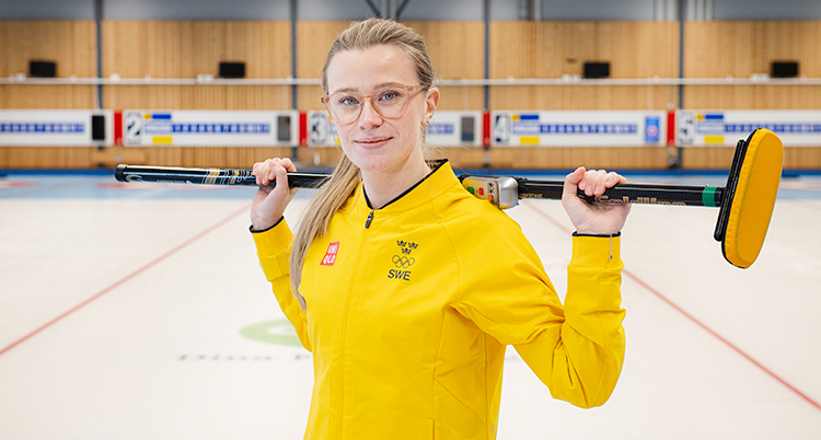 Hon står på en is för curling. Hon tittar in i curling. Hon håller en sop för curling över sina axlar.