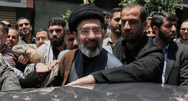 Han står vid en bil. Det är fullt med män runt honom. Han har glasögon och en turban på huvudet.