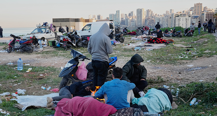 Människor sitter på marken. De har med sig väskor och filtar. I bakgrunden syns staden Beirut.