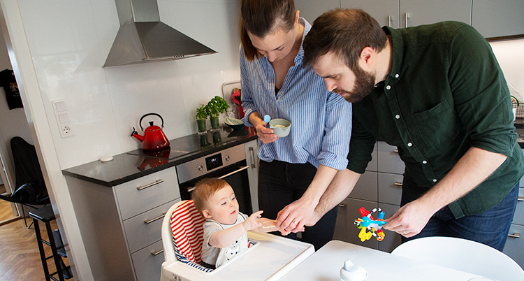 Ett kök. En kvinna och en man står vid bordet. En bebis sitter i en barnstol.