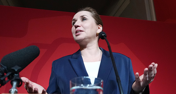 Hon pratar på enscen. Hon håller ut armarna. Bakom henne finns ett rött skynke.