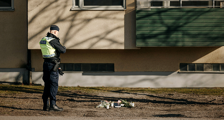 En polis står framför ett hus med lägenheter. På marken ligger blommor.