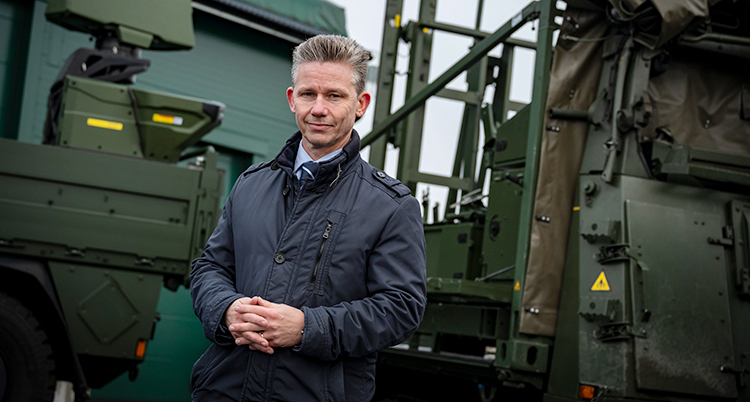 Han står bland några militära fordon. Han tittar in i kameran.