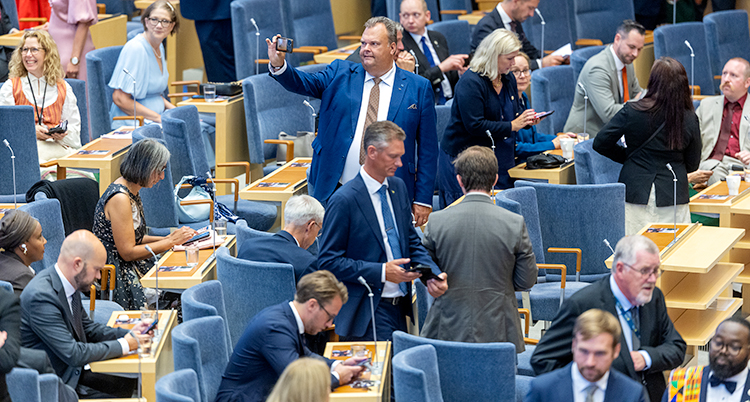 Riksdagens stora sal. Det är paus. Politiker står eller sitter och pratar med varandra.