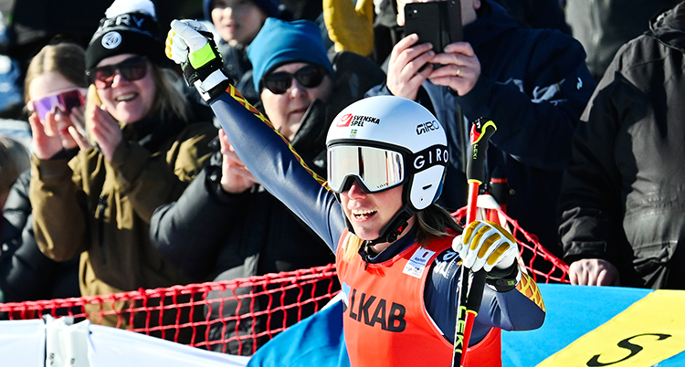 Hon är glad. Hon sträcker upp en arm. Hon har hjälm och skidglasögon.