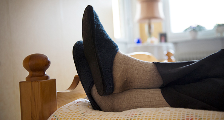 En person ligger på en säng. Närbild på personens ben. Personen har tofflor på sig.