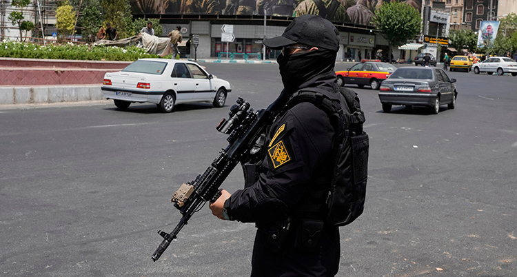 Han håller i ett gevär. Han står vid en vägg. Bilar åker förbi honom.