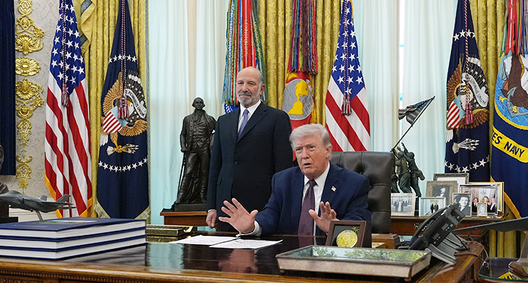 Donald Trump sitter bakom ett skrivbord i Vita huset. Bakom honom står en man som jobbar åt honom.