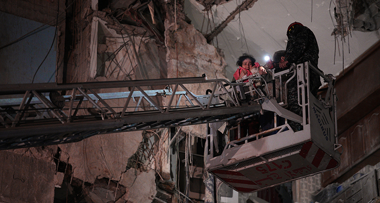 Det är mörkt ute. En kvinna får åka i en korg som sitter på en lyftkran. I bakgrunden syns ett hus som är förstört av bomber.
