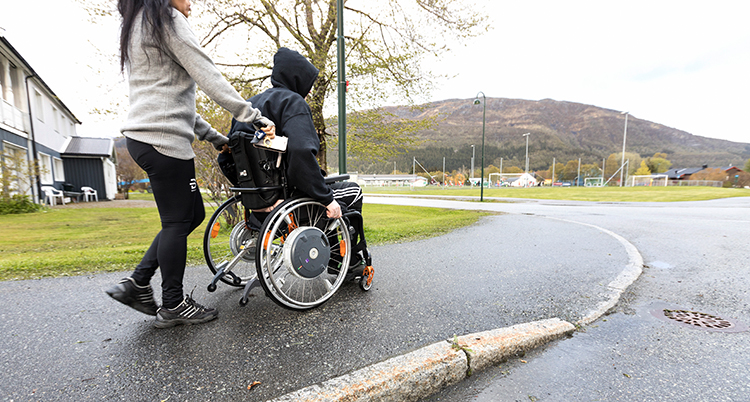 En kvinna går på trottoaren och skjuter en rullstol framför sig. I rullstolen sitter en person.