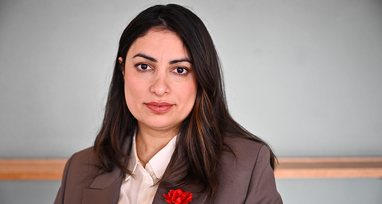 En närbild på henne. Hon har mörkt långt hår. Hon tittar in i kameran. Hon har skjorta och kavaj och en röd blomma på kavajslaget.