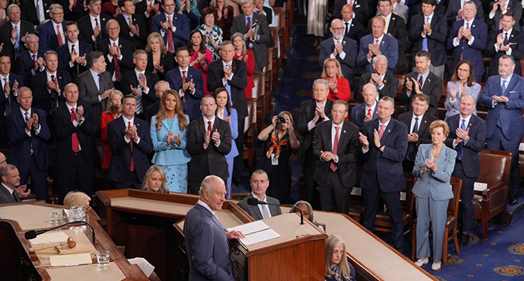 Charles står i en talarstol med en mikrofon. Politikerna står upp och applåderar.