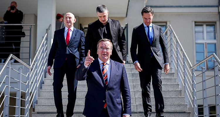 Han har kostym och pekar med sitt finger. Bakom honom syns tre andra män i kostym.