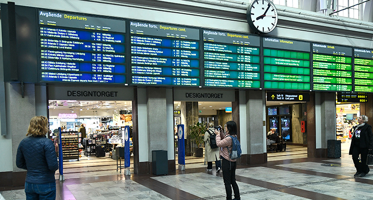 Människor står på stationen framför en tavla där det står om alla resor.