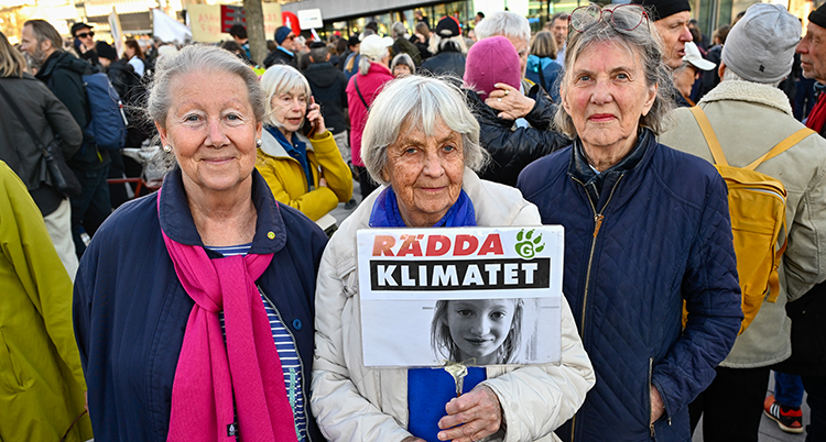 Tre äldre damer tittar in i kameran. Kvinnan i mitten håller upp en skylt med texten Rädda klimatet.