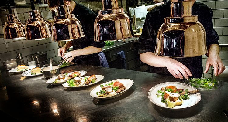 Två kockar som jobbar i ett kök på en restaurang. Kockarna är män. De lägger upp mat på flera tallrikar.
