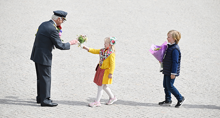 Kungen står på ett torg. Två barn går fram till kungen med blommor.