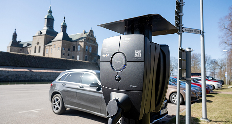 En solig dag. Närmast i bild är en laddstolpe. Bakom en parkering med bilar. Och längst bak Vadstena slott.