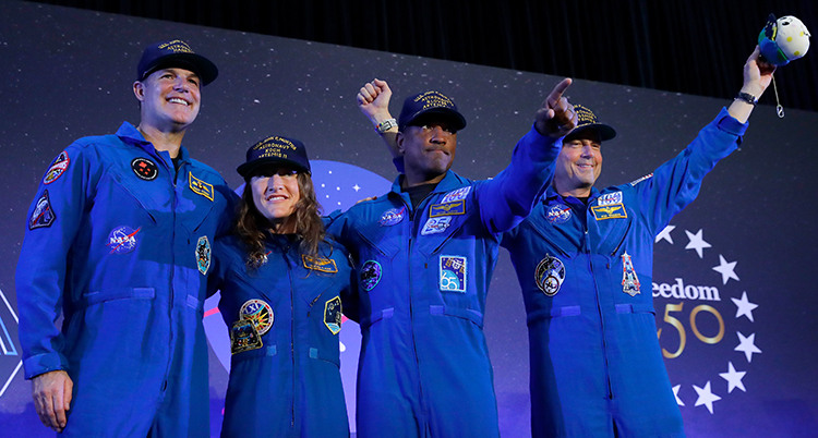 De står på en rad. De har blåa overaller och kepsar. De är glada och viftar med armarna.