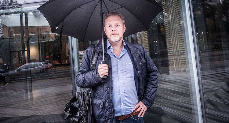 Han står med ett paraply. Han tittar in i kameran.