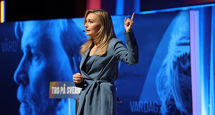 Hon står på en scen. Hon pekar med ett finger.