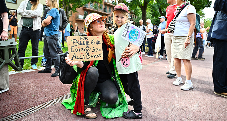Mamman sitter på knä bredvid dottern. De håller i en skylt.