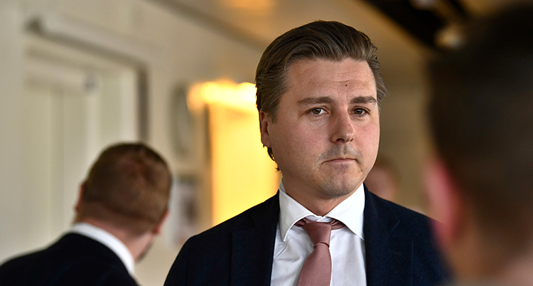 Han har kostym och slips. Han står i en korridor. En man går förbi.