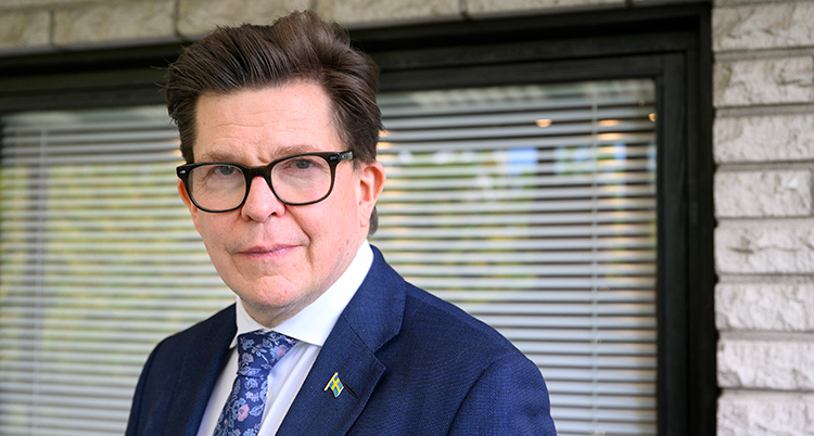 Han står framför ett fönster. Han har kostym, slips och glasögon.