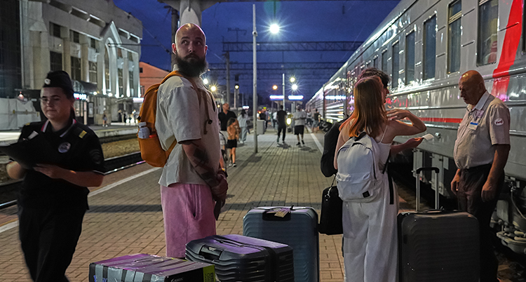 En lång man står på en station. Han har fler väskor. Runt honom står andra personer. Ett tåg har stannat vid stationen.