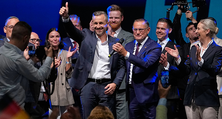 En grupp människor ser glada ut. Mannen i mitten längst fram sträcker upp armen och gör en tummen upp.