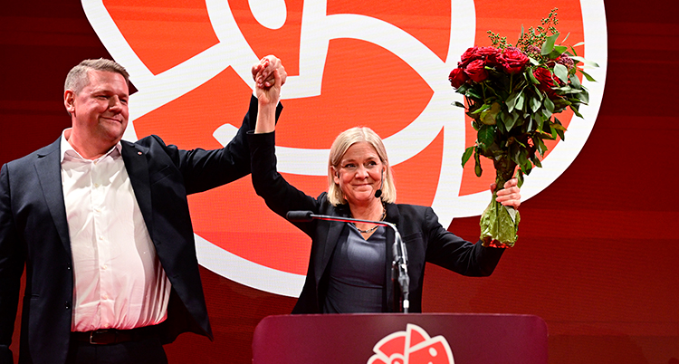 Magdalena Andersson och en person i hennes parti sträcker upp armarna till seger. Andersson håller en blombukett.