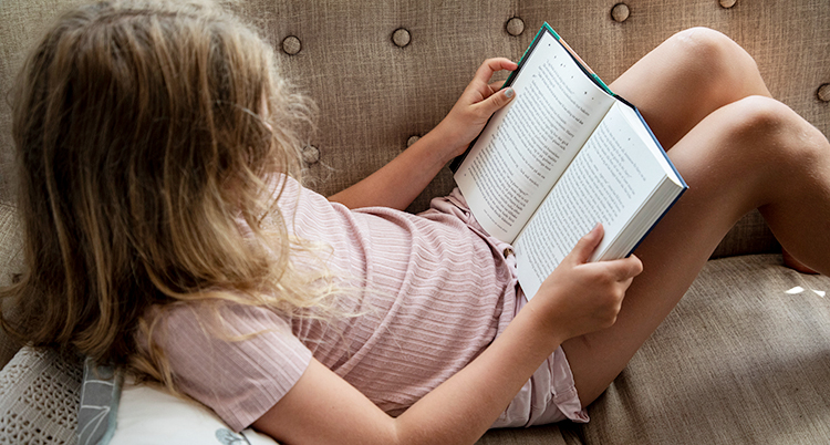 En flicka ligger i en soffa och läser en bok.