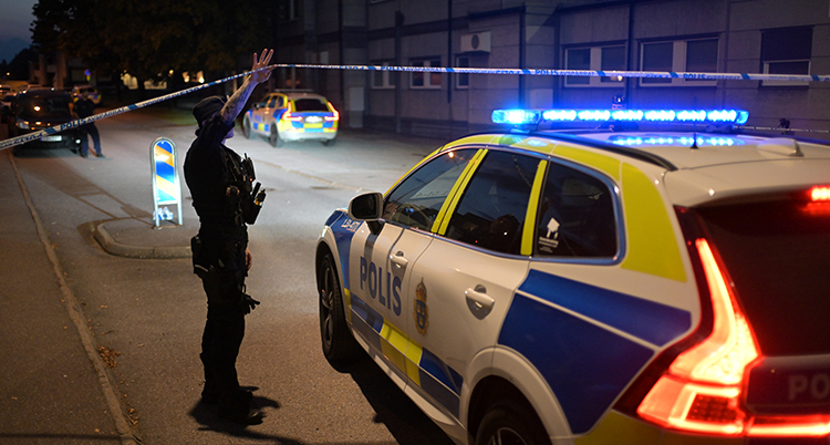 Poliser är vid en mörk plats. En polisbil är parkerad. I bakgrunden är det hus med lägenheter.