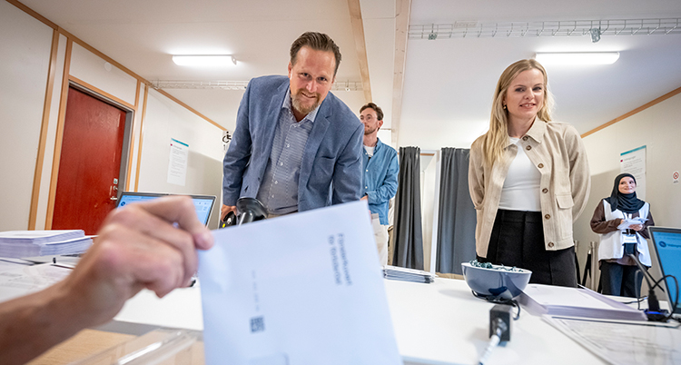 En hand lägger ner ett kuvert i lådan. Bakom syns en man och en kvinna som har röstat.