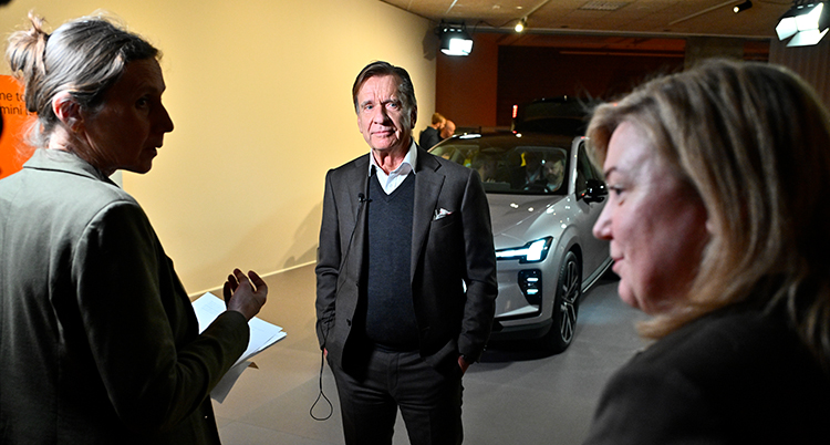 Han står framför en bil. Han pratar i mikrofoner som journalister håller fram.