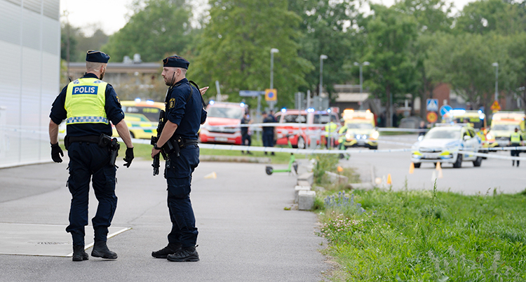 Två poliser står utomhus. Längre bort finns polisbilar och ambulanser.