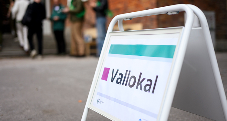 En skylt står utomhus. På skylten står det Vallokal. Bakom syns några personer som köar vid ett hus.