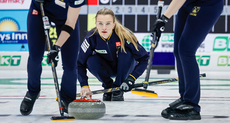 Hon sitter på isen efter att ha släppt iväg curlingstenen. Två andra spelare sopar framför stenen.